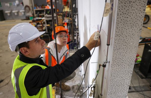 Dr Enrique del Rey Castillo and PhD student Victor Li both wearing hard hats and high-vis jackets, attach sensors to a vertical wall in a warehouse.