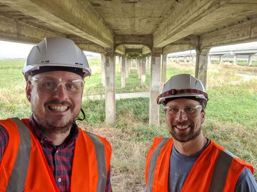 Dr Lucas Hogan and Dr Max Stephens, wearing hard hats and hi-vis jackets, stand smiling at the camera under a bridge.