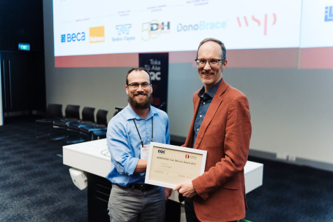 Dr Lucas Hogan and Professor Ken Elwood looking at the camera, smiling, shaking hands and holding Dr Lucas Hogan's certificate.  