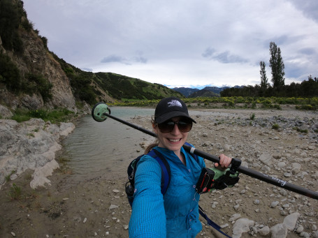 Erin McEwan, in a coat and cap with a walking pole over her shoulder, smiles at the camera. She is stood near a river during fieldwork at the Waiau Toa/Clarence River Valley.